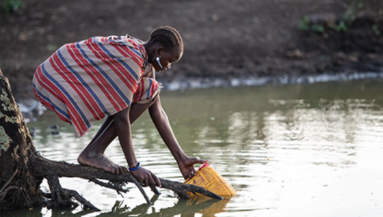 A South Sudanese woman crouching down to collect water from a natural body of water